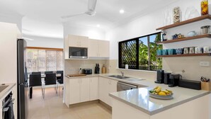 A modern kitchen with white cabinets, a microwave, a toaster, and a sink. There is a dining area with a table and chairs, and a window with a view of greenery.