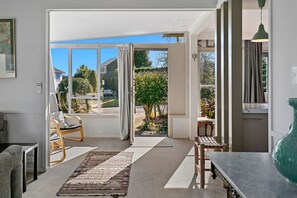 A modern living room with a large sliding glass door leading to a garden. There are two wooden chairs, a small table, and a rug on the floor.