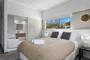 A modern bedroom with a large bed, a bathroom with a mirror and sink, and a window with a view of trees and a house. - "The Maple Tree" Taupo - central accommodation (Taupo)