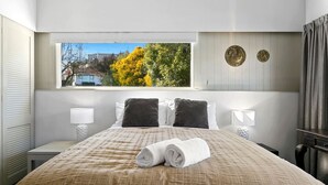 Habitación de hotel con una cama grande, dos almohadas, un ventanal con vistas a árboles y una mesita de noche con lámpara.