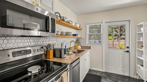 Eine moderne Küche mit Edelstahlgeräten, einer Mikrowelle und einem Toaster. Über der Arbeitsplatte befinden sich Regale mit Geschirr, und ein Fenster mit Jalousien.