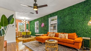 Ein Zimmer mit grüner Wand, Holzboden, orangefarbenem Sofa, rundem Korbstuhl-Couchtisch und Deckenventilator.