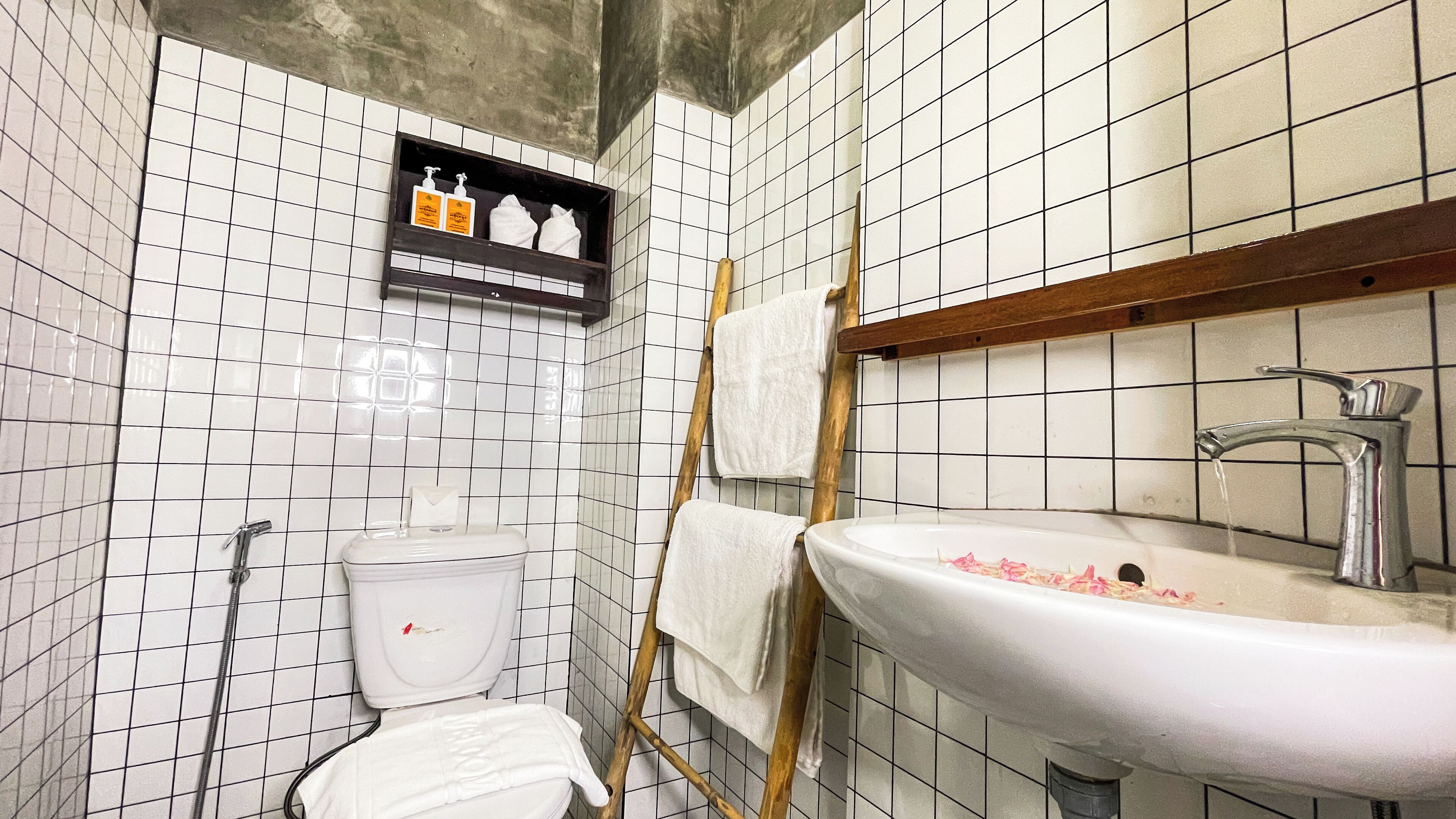 Een badkamer met witte betegelde wanden, een toilet, een wastafel met kraan en een plank met toiletartikelen.