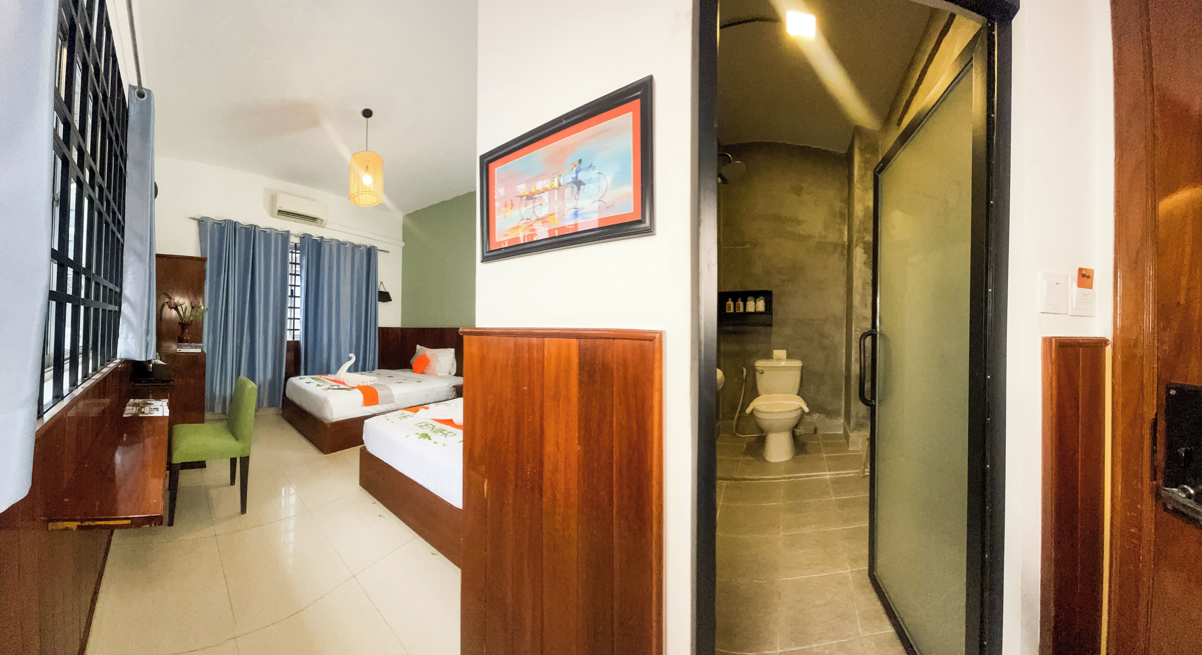 Una camera d'albergo con una scrivania in legno, una sedia verde, un letto con copriletto bianco e arancione, un quadro incorniciato appeso al muro e un bagno con il water e la doccia.