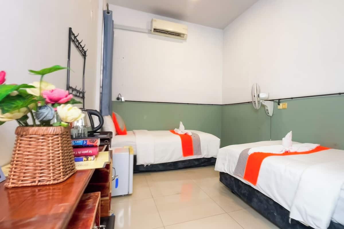 A hotel room with two beds, a small desk with a basket of flowers, a bookshelf, and a wall-mounted air conditioner.