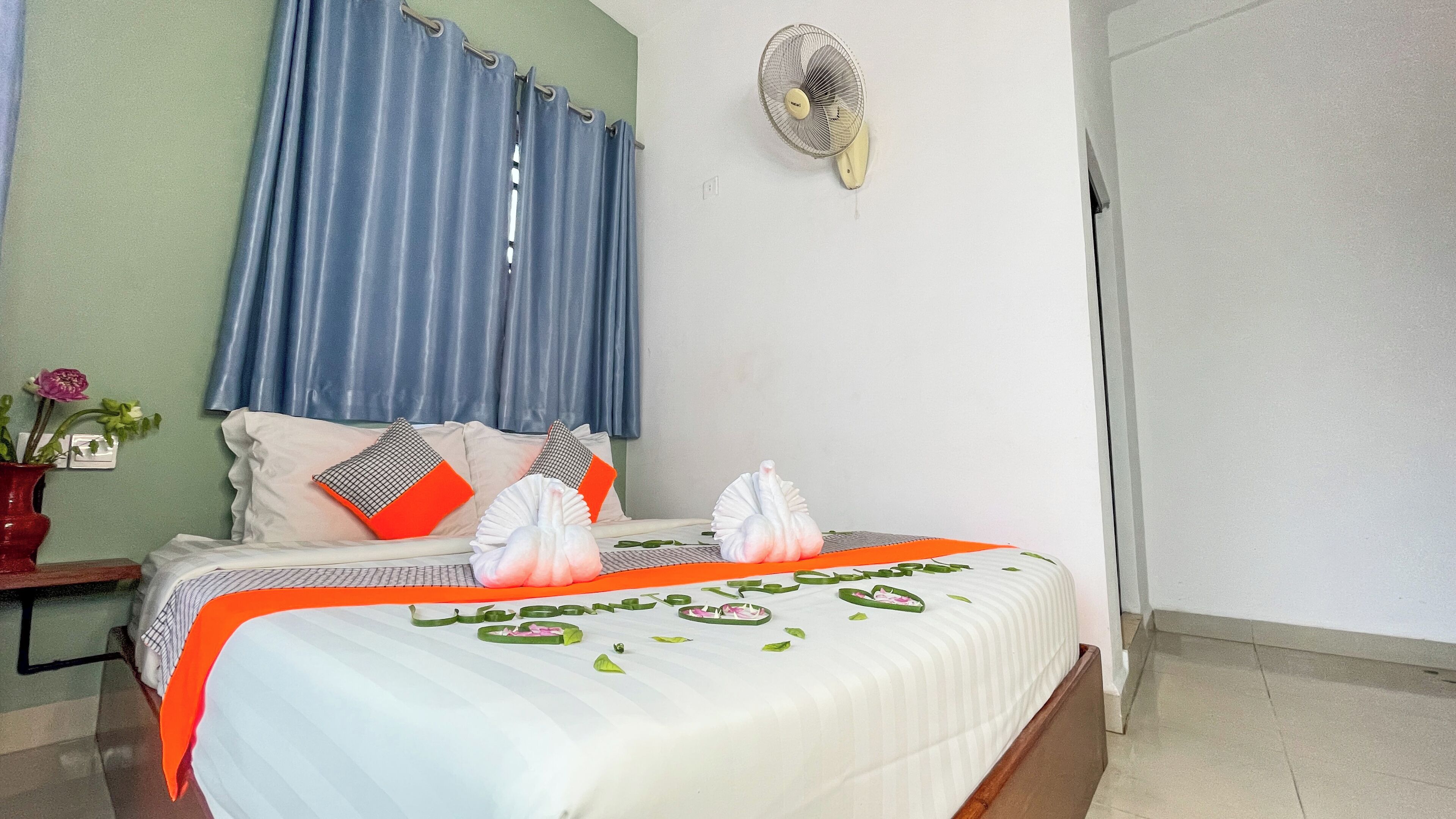 Cama bem arrumada com colcha branca e laranja, dois travesseiros e cisnes decorativos. Um ventilador de parede está montado na parede acima da cama.