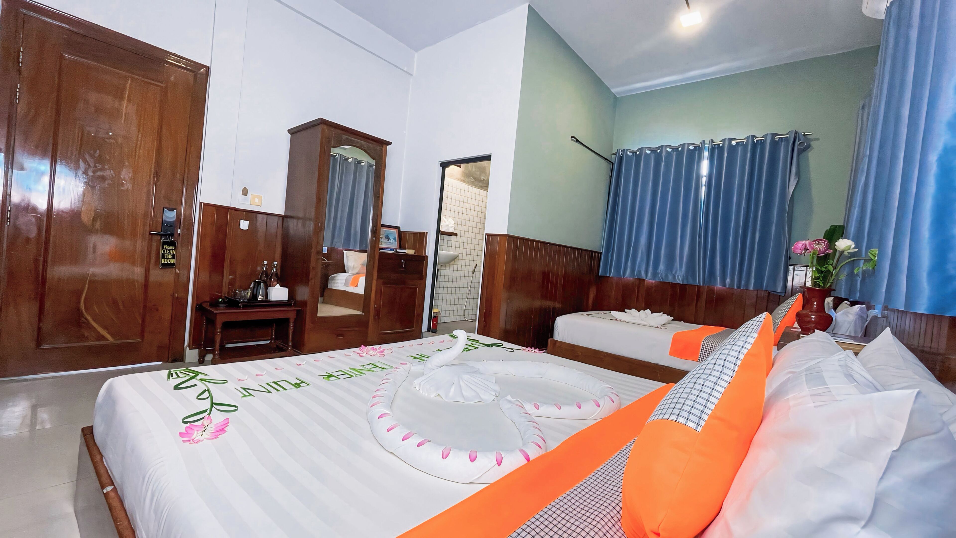 Habitación de hotel con una cama grande, una cama más pequeña, un armario de madera, un escritorio con una silla y un baño visible a través de una puerta abierta.