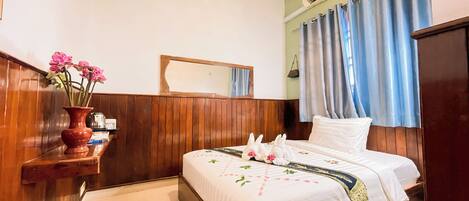 A hotel room with a wooden headboard, a bed with a white and blue bedspread, a vase with pink flowers on a wooden shelf, and a mirror on the wall.