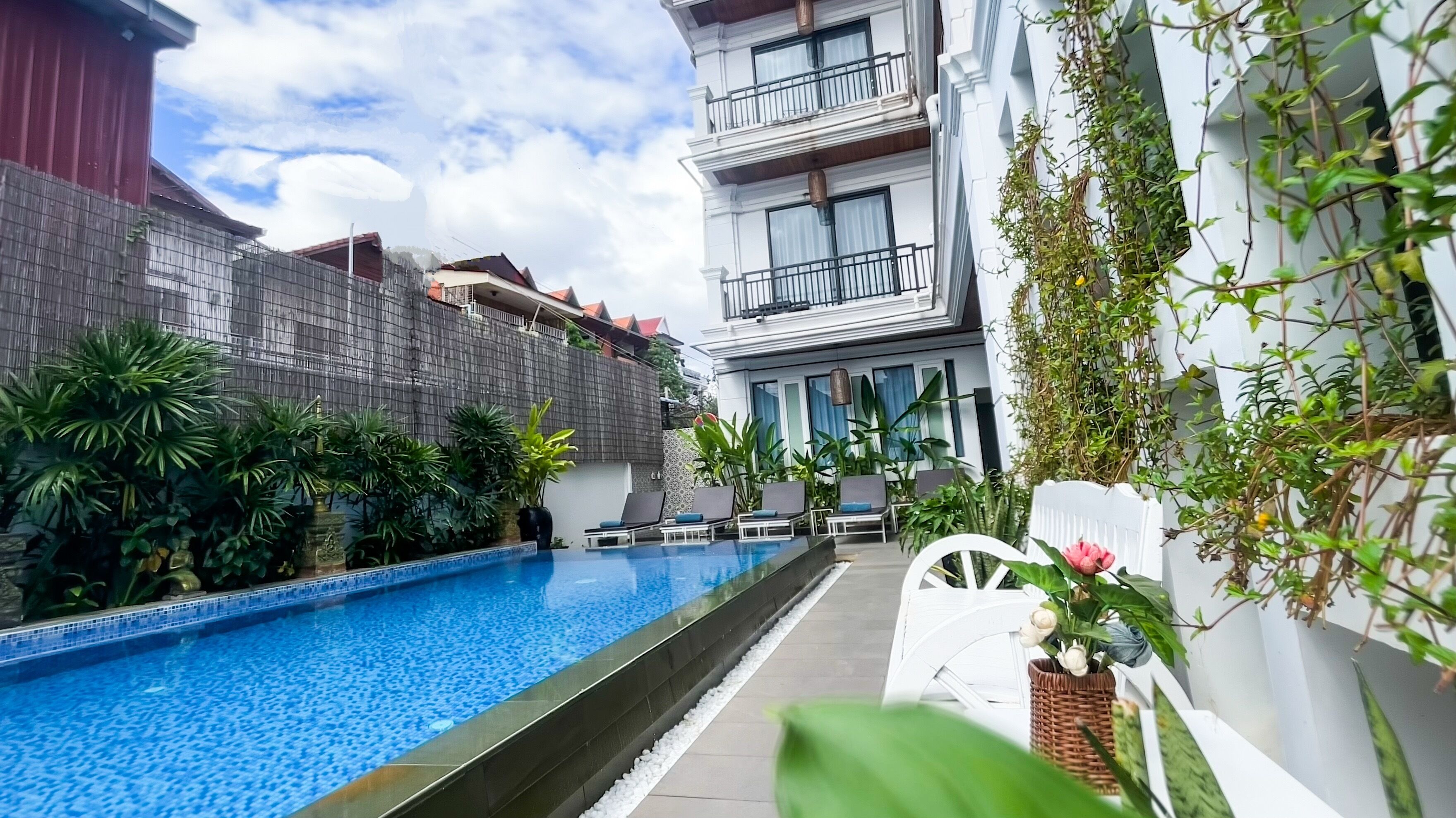 Piscina con sillas de descanso y plantas en macetas, rodeada por un edificio con balcones y una valla.