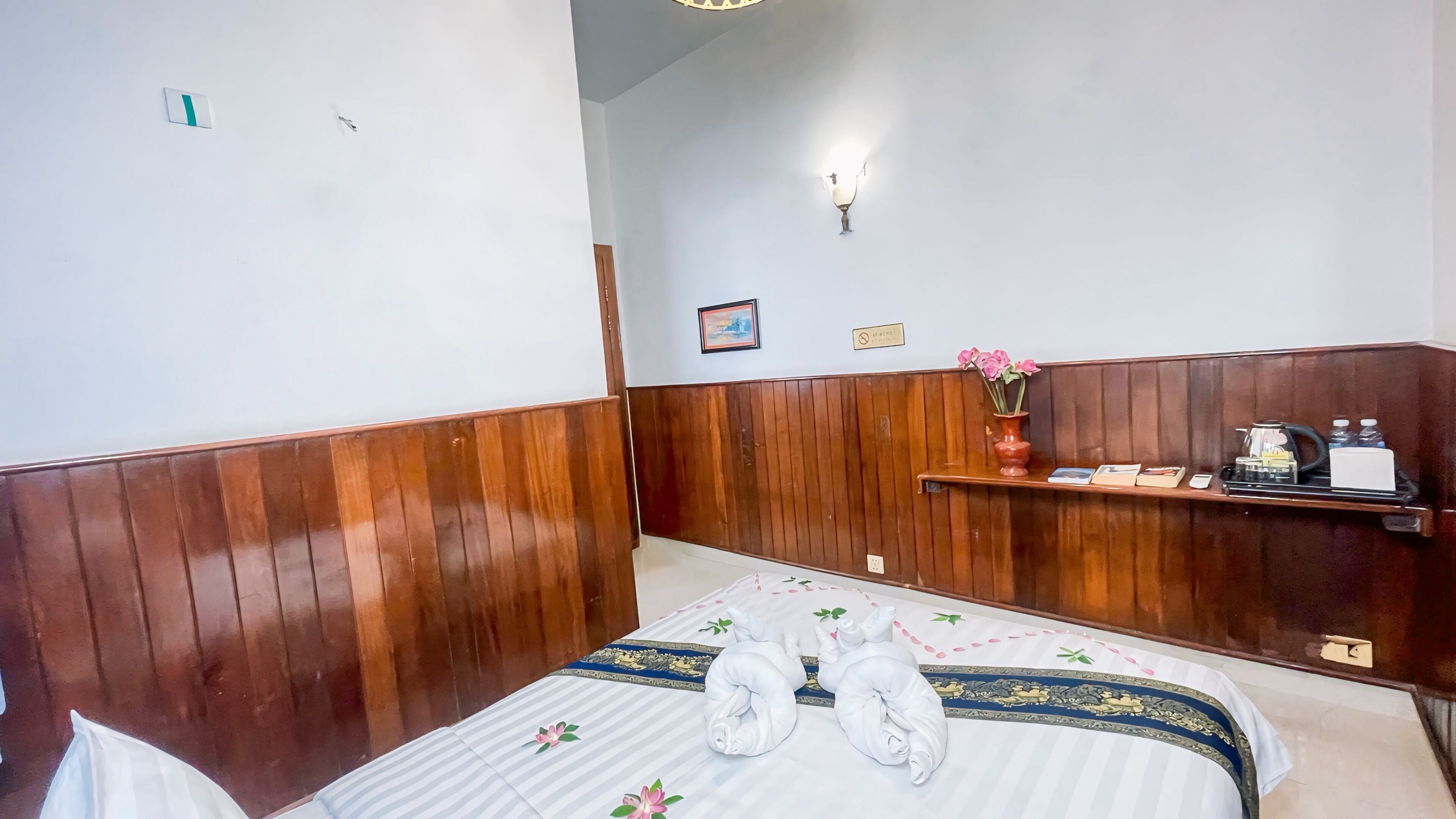 Une chambre d’hôtel avec un mur lambrissé en bois, un lit avec du linge de lit et des serviettes blanches, une petite étagère ornée d’un vase de fleurs et un tableau encadré accroché au mur.
