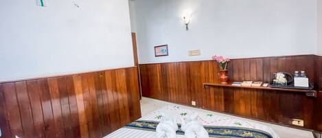Habitación de hotel con pared de paneles de madera, cama con ropa blanca y toallas, una pequeña estantería con un jarrón de flores y un cuadro en la pared.