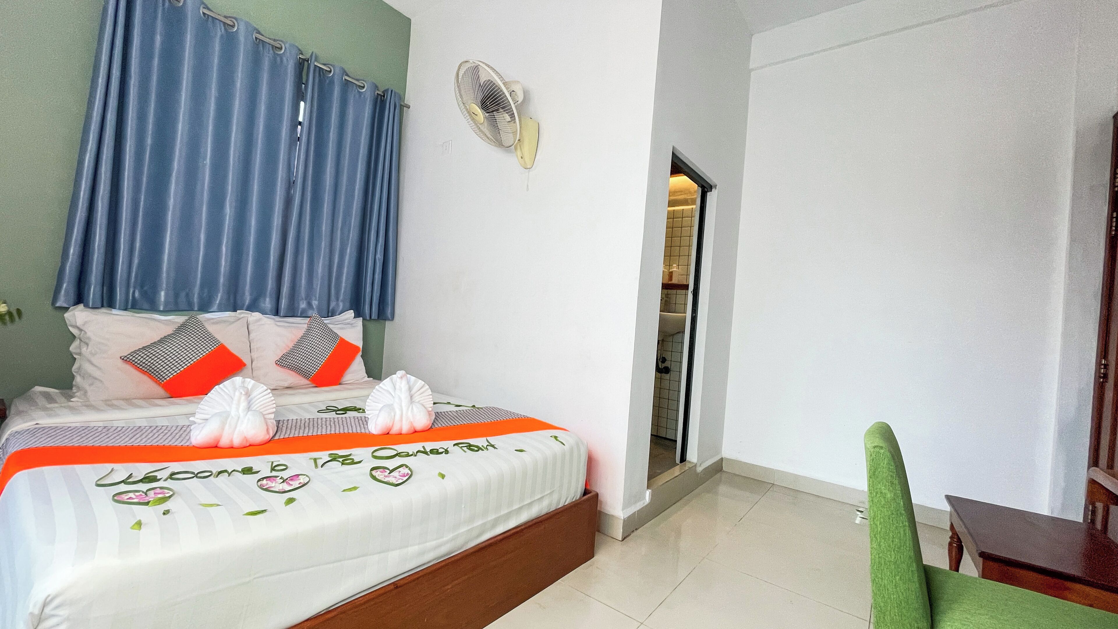 A hotel room with a bed, a fan, a green chair, and a door leading to a bathroom.