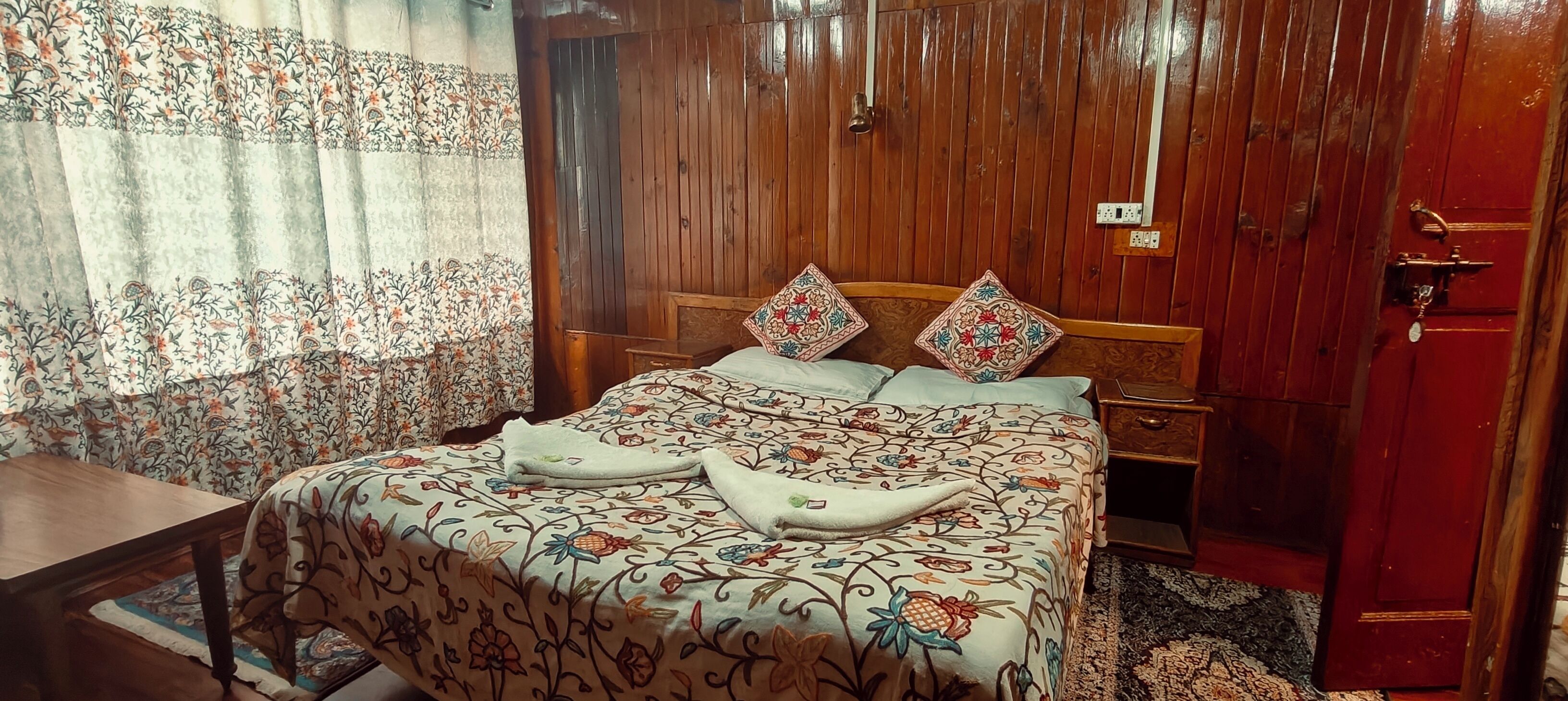 A bedroom with a wooden headboard, floral bedspread, two pillows, a bedside table, and a wooden wardrobe.
