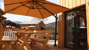 Une terrasse en bois avec un grand parasol, des bancs en bois et une porte vitrée donnant sur une pièce.