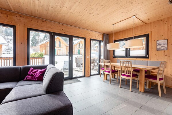 A modern interior with a wooden ceiling, a grey sofa with a purple pillow, a wooden dining table with chairs, and large windows offering a view of wooden cabins outside.