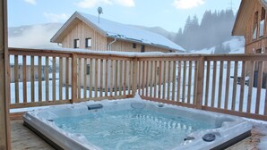 Eine Holzterrasse mit Whirlpool, umgeben von schneebedeckten Gebäuden und Bergen im Hintergrund.