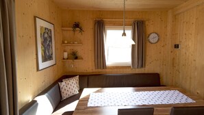 A cozy cabin interior with a wooden dining table, cushioned seating, a clock, and a window with curtains.