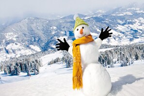Un muñeco de nieve con sombrero verde y bufanda amarilla en un paisaje nevado con montañas al fondo.