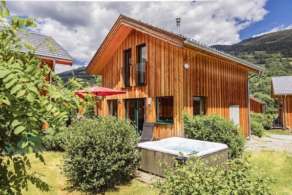 A wooden cabin with a hot tub in the yard, surrounded by greenery and mountains in the background.