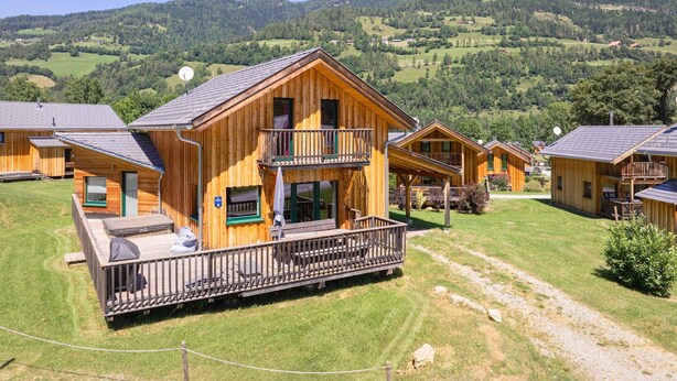 A wooden cabin with a balcony, surrounded by a grassy area and a mountainous landscape.