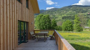 Un edificio de madera con terraza, asientos exteriores y vistas a colinas verdes y una montaña.