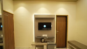 Ein Hotelzimmer mit Flachbildfernseher an der Wand, einem kleinen Tisch mit Wasserkocher, einer Bank und einem Kühlschrank.