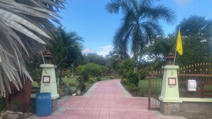 A paved pathway leading to a gated entrance with a yellow flag, surrounded by lush greenery and palm trees. - House for rent in Hua Hin for wintering 180 days visa (Hua Hin)
