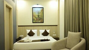 A hotel room with a bed, a chair, a bedside table, a nightstand, a framed picture on the wall, and a window with curtains.