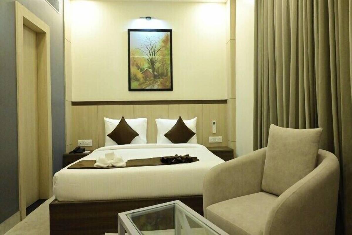 A hotel room with a bed, a chair, a bedside table, a nightstand, a framed picture on the wall, and a window with curtains.