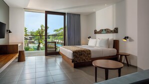 A modern hotel room with a large bed, a desk, a small table, and a balcony with a view of greenery and buildings. - Dialoog Seminyak (Keroboken Kelod)