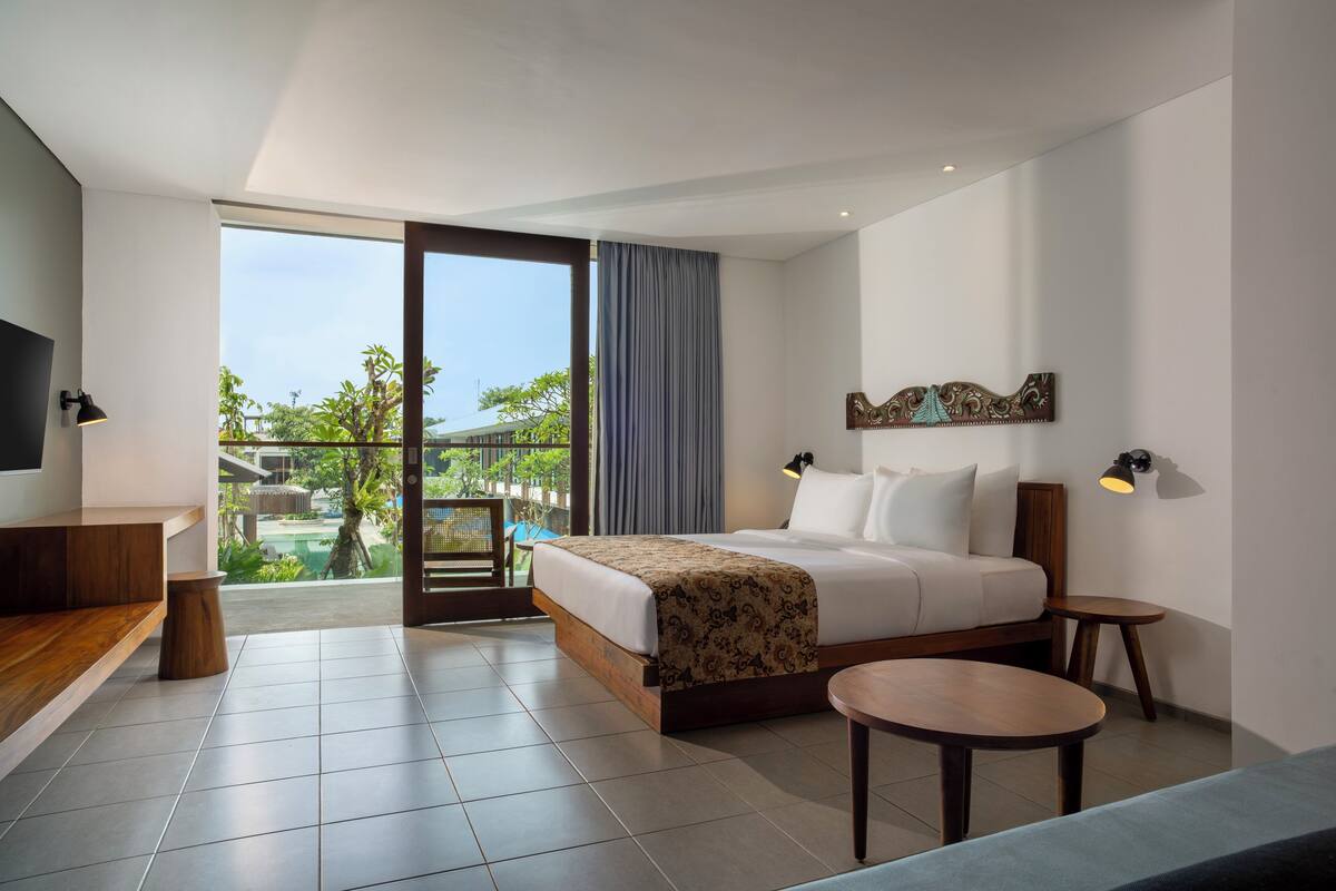 A modern hotel room with a large bed, a desk, a small table, and a balcony with a view of greenery and buildings.