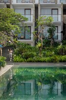 Un espace piscine où l’on peut admirer le reflet du bâtiment, entouré d’une végétation luxuriante, et où une personne se promène.
