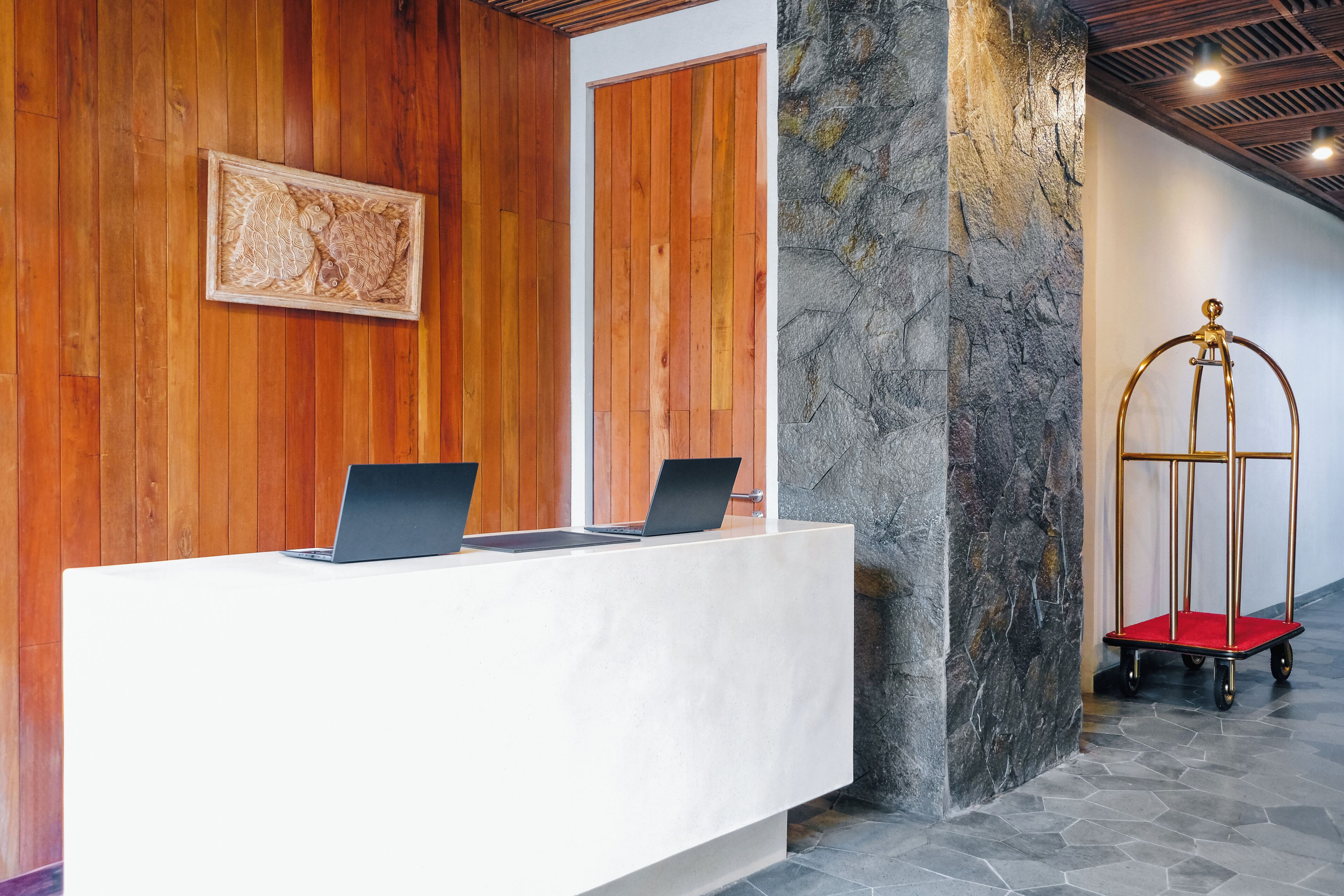 Un mostrador de recepción con dos computadoras portátiles, un panel de madera, un pilar de piedra y un carrito con marco dorado.