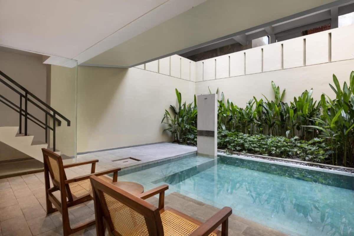 A swimming pool with a glass barrier, surrounded by plants and wooden chairs.