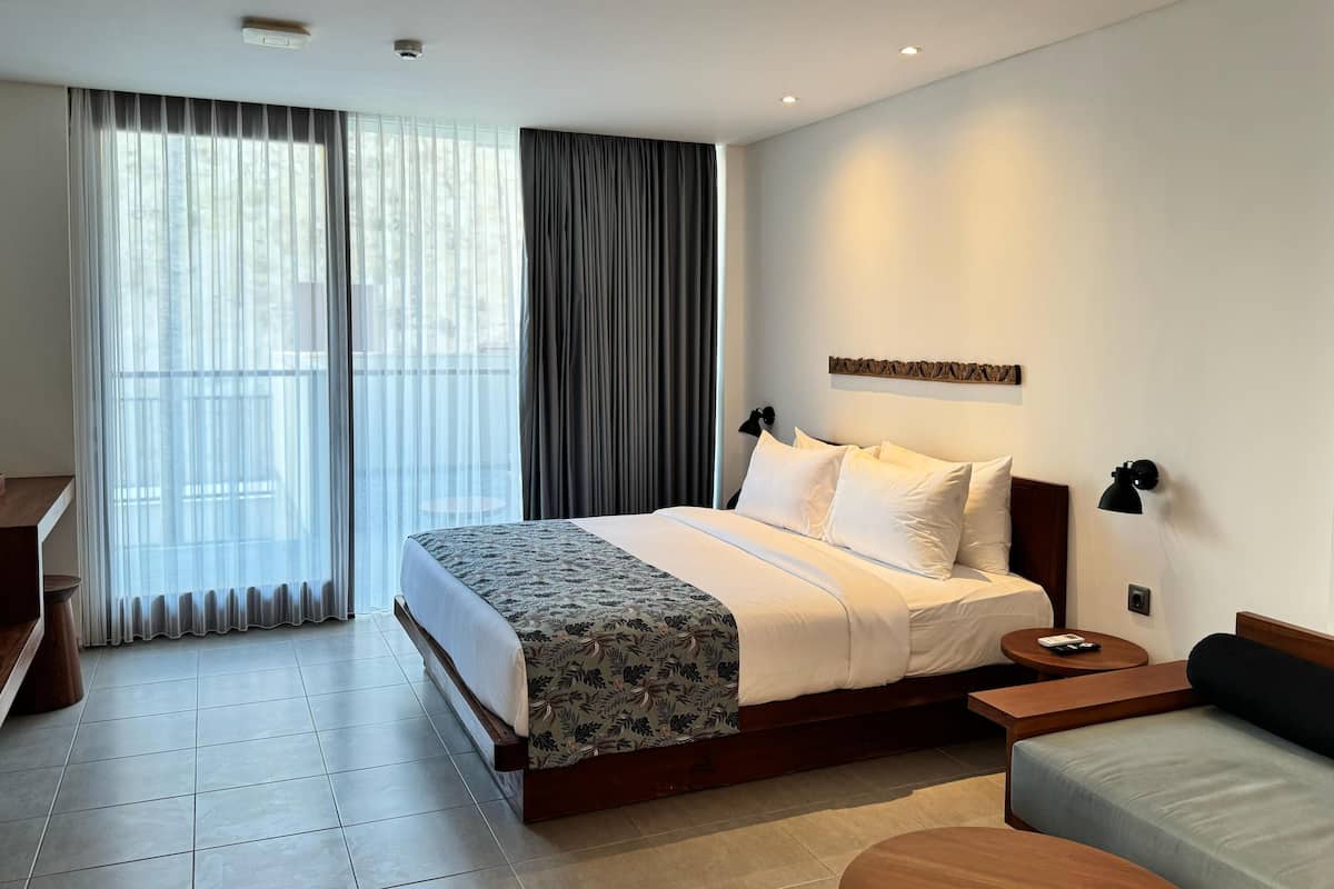 A modern hotel room with a large bed, a bedside table, a small sofa, and a view of the outdoors through sheer curtains.