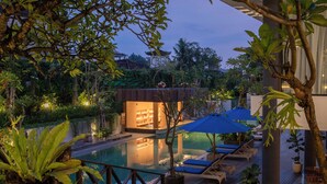 A pool area with lounge chairs and umbrellas, surrounded by lush greenery and a building with a modern design. - Dialoog Seminyak (Keroboken Kelod)