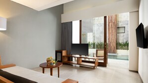 A modern living room with a flat-screen TV, wooden furniture, and a large window with a view of a pool and greenery. - Dialoog Seminyak (Keroboken Kelod)