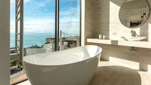 Un baño moderno con una bañera independiente, un espejo redondo y vistas al mar a través de amplios ventanales.