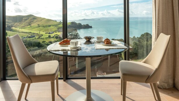 Un comedor con una mesa de vidrio y dos sillas, con vistas a un paisaje pintoresco con colinas y una masa de agua.