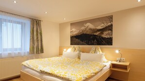 A bedroom with a large bed, a nightstand with a lamp, a window with sheer curtains, and a mountain landscape painting on the wall. - Cozy vacation apartment and perfect base in the heart of Tyrol (Inzing)