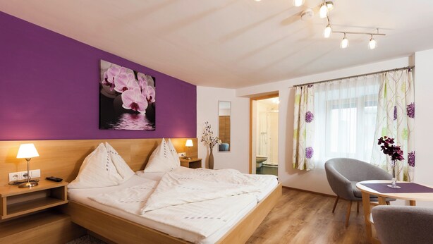 A hotel room with a bed, nightstands, a chair, a table, and a bathroom visible through an open door. - Cozy vacation apartment and perfect base in the heart of Tyrol (Inzing)
