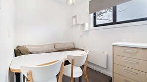 A compact, modern room with a white round table and chairs, a beige sofa, a window with a view of trees, and a wooden cabinet with drawers. - Celesia 2 - Central Village (Central Village)