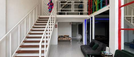 Una escalera conduce a un balcón en el segundo piso, con barandilla de colores, un sofá verde y un ventilador en el suelo.