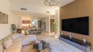 Een moderne woonkamer met een grote flatscreen-tv, een bank, een salontafel en een eethoek op de achtergrond.