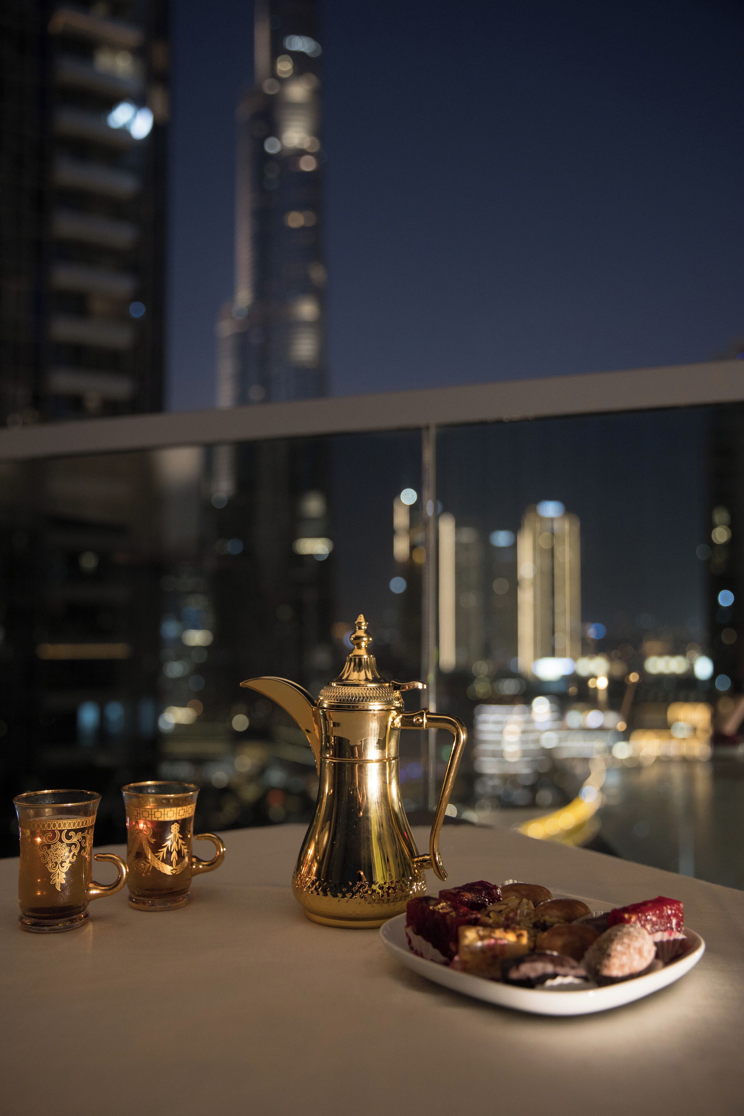 A table with a golden teapot, two ornate cups, and a plate of food against a cityscape backdrop.