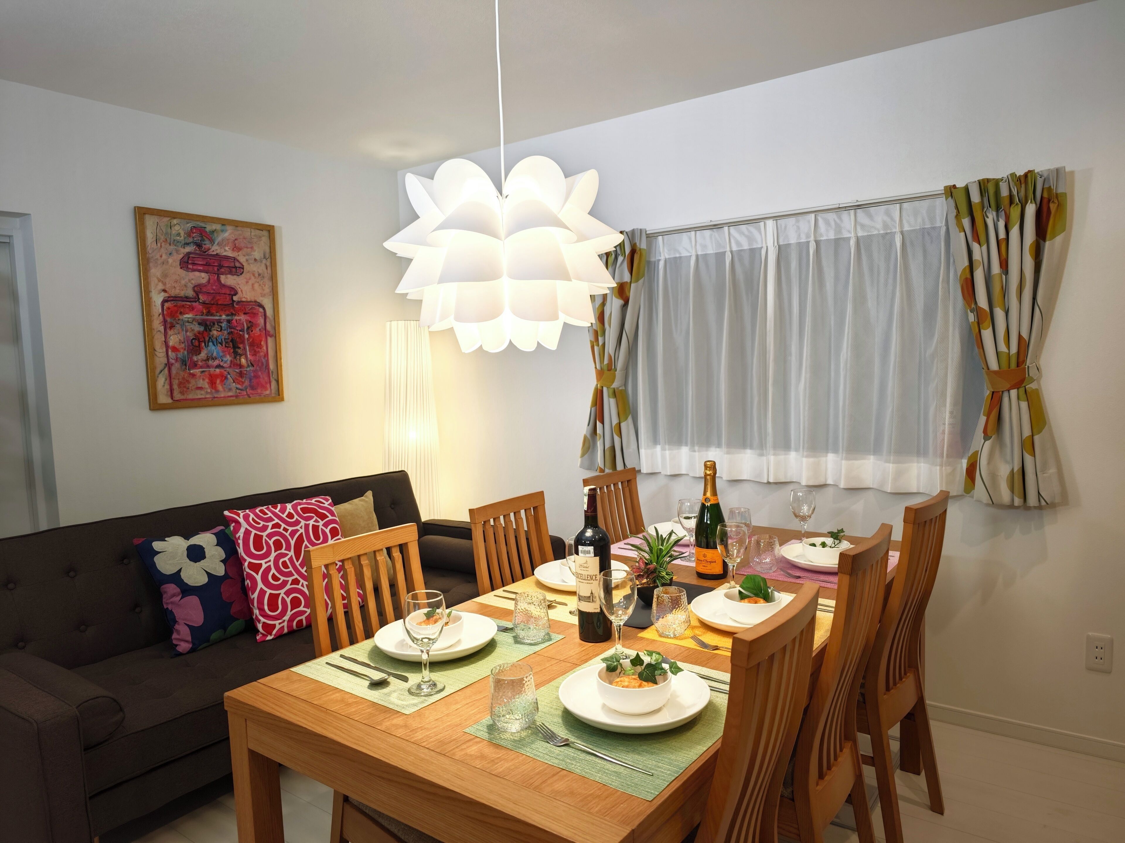 Ein Essbereich mit einem Holztisch, gedeckt für zwei Personen, umgeben von Holzstühlen. Eine Chandelüster hängt über dem Tisch, und im Hintergrund ist ein Fenster mit Vorhängen zu sehen.