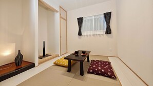 A minimalist living room with tatami flooring, a low table with cushions, a lamp, and a window with curtains. - K-icon Hotels Chikusa (Nagoya)