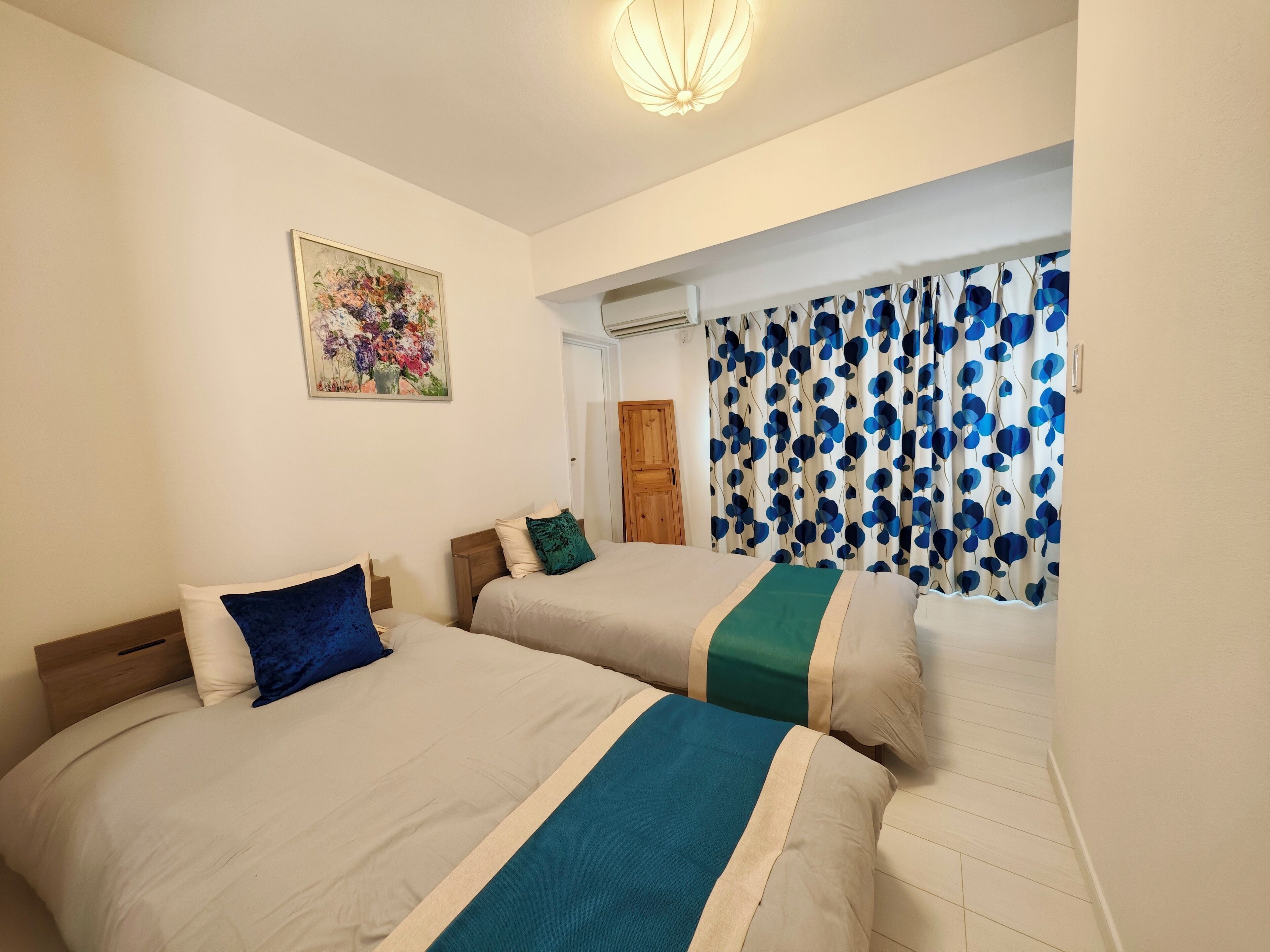 Habitación de hotel con dos camas, un cuadro floral en la pared y una ventana con cortinas azules y blancas.