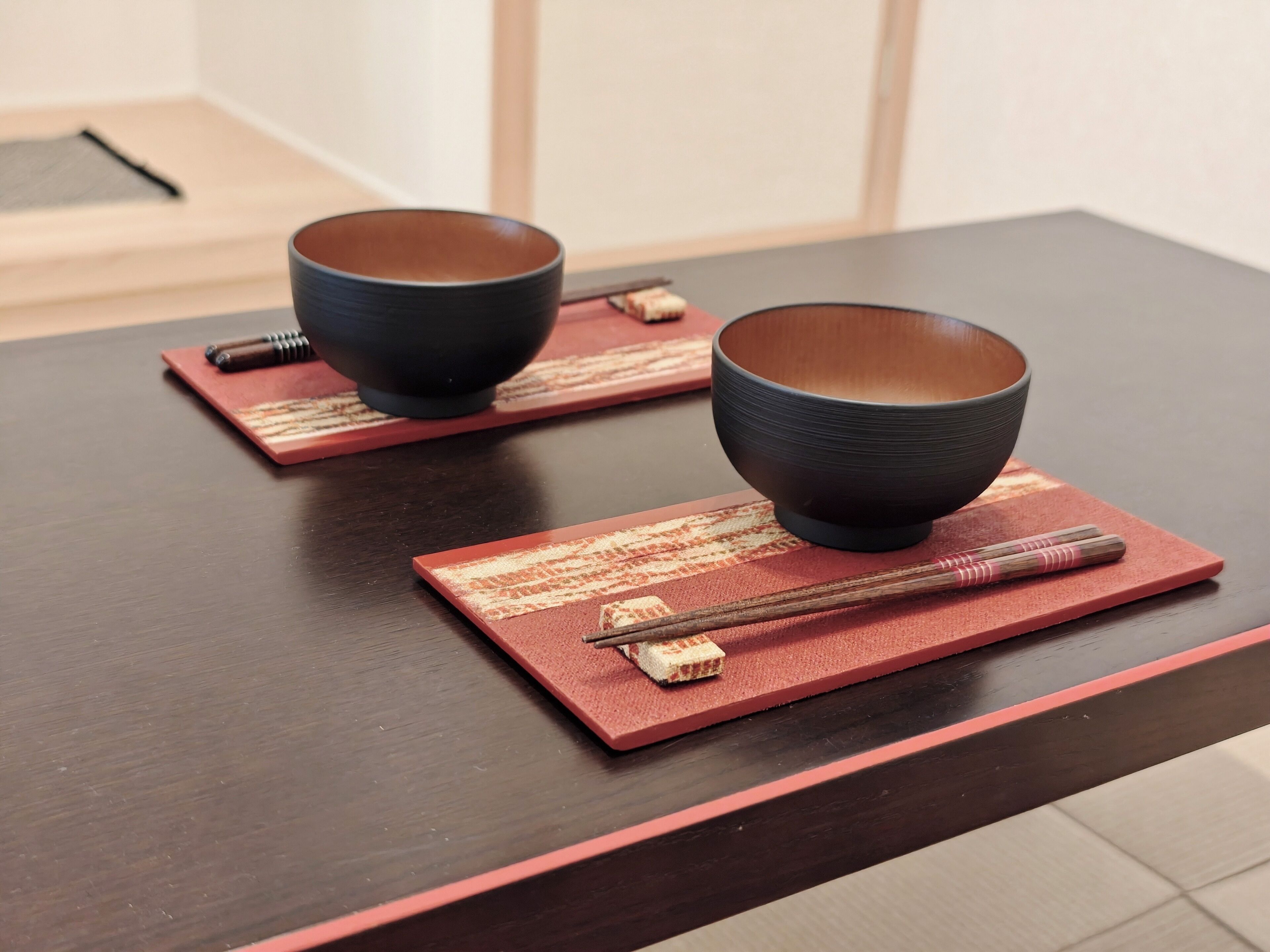 Two bowls on a dark wooden table with chopsticks and a red placemat.