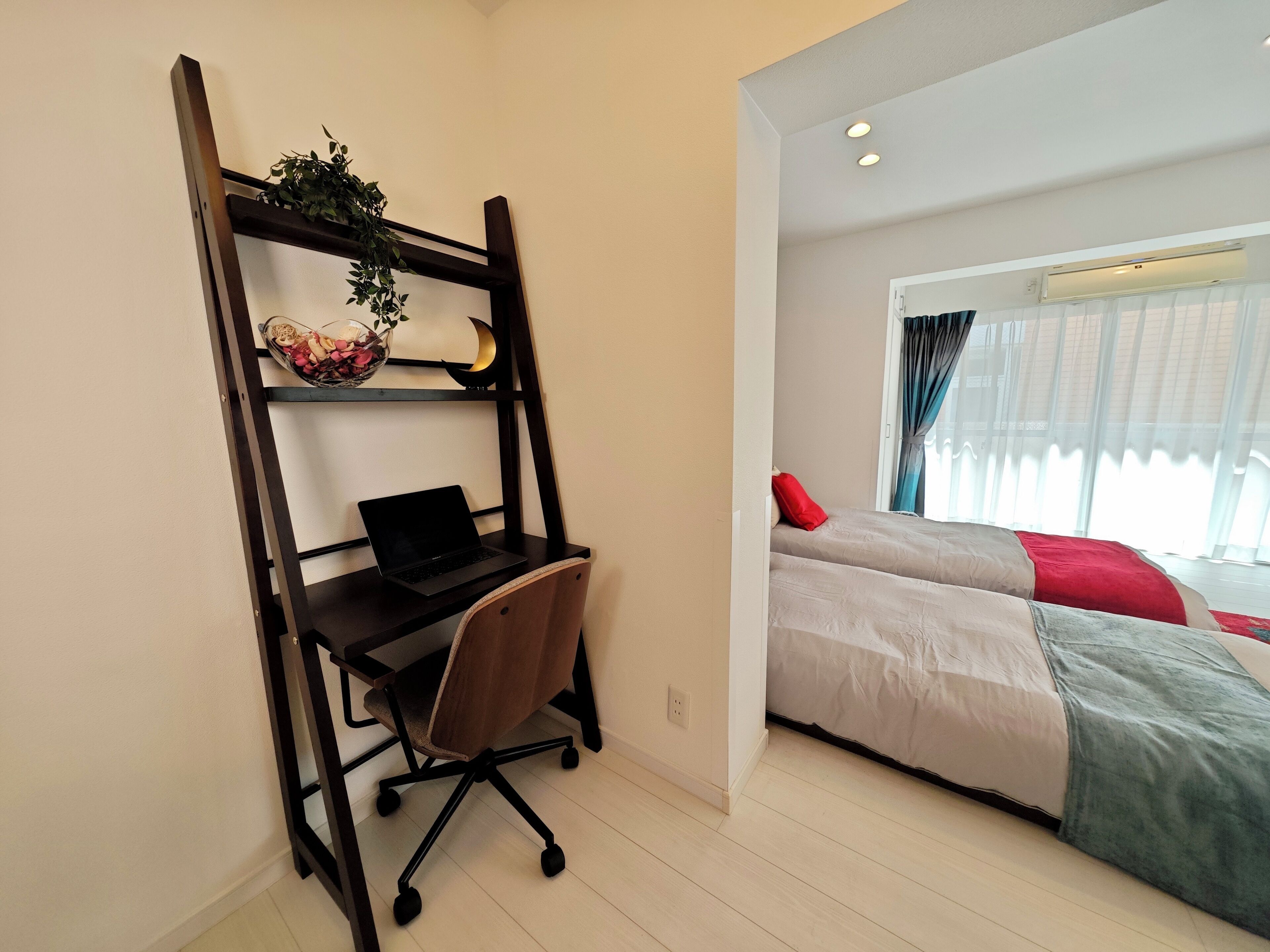 A bedroom with a bed, a desk with a laptop, and a shelf with a plant and decorative items.